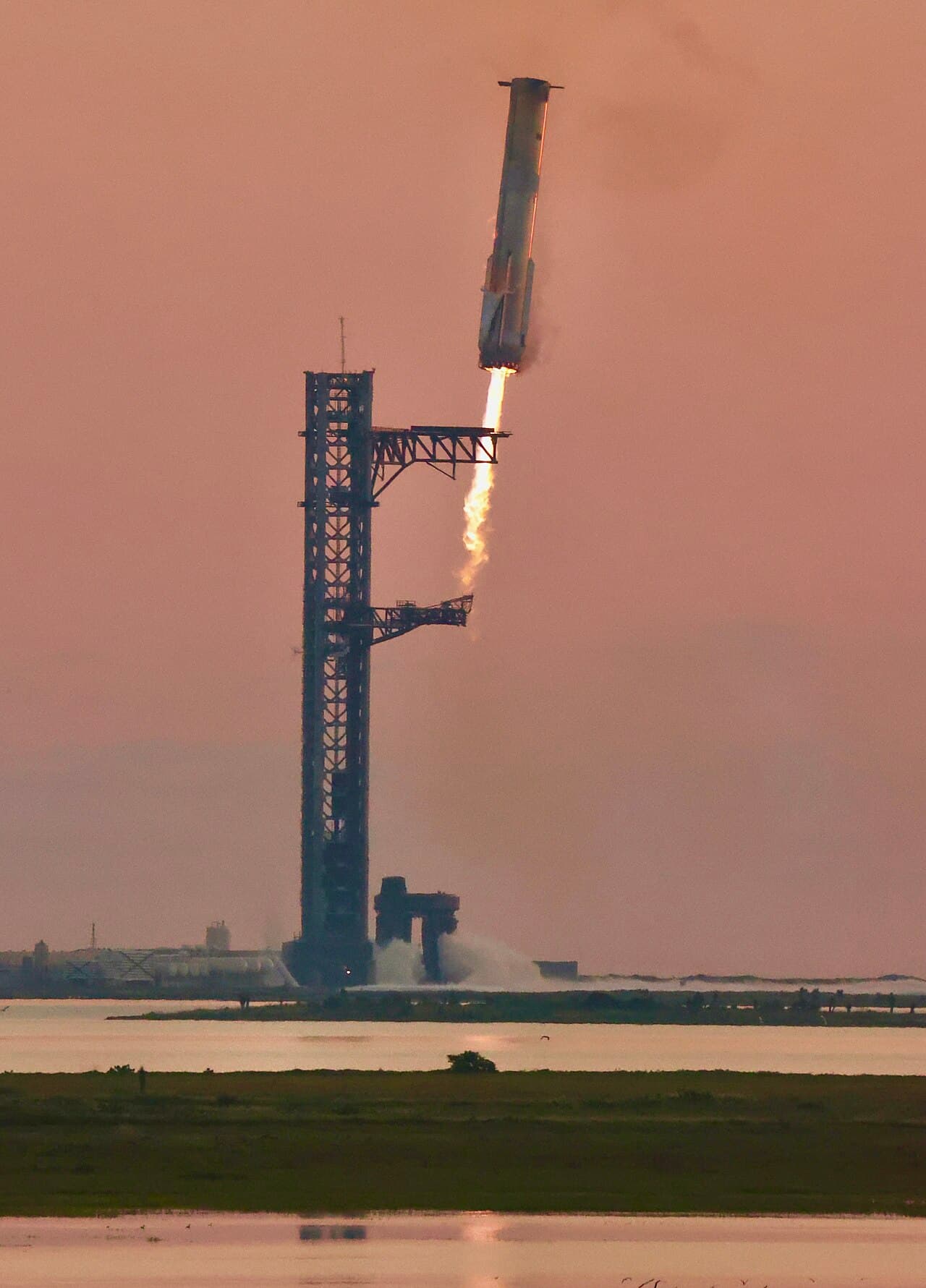 The Super Heavy booster descending vertically alongside the launch tower on final approach, engines lit.